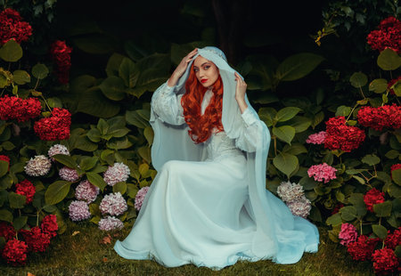 Fantasy woman in white vintage dress head covered with veil. Red-haired girl queen Medieval princess bride sits in garden green trees bushes hydrangea flowers. Mystical image sexy lady gothic vampireの写真素材