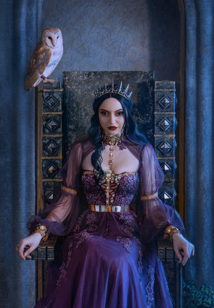 Mythical fantasy woman queen vamp sits on medieval ancient throne. Golden gothic crown on head. Elf girl princess evil face black long hair. Purple vintage art dress. Barn Owl bird symbol wisdomの写真素材