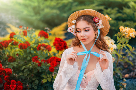 Fantasy girl princess beautiful face, delicate make-up sttraw hat on head, long hair blue ribbon. Happy woman sits in garden enjoys nature green bushes red flowers roses. art pink white vintage dressの写真素材