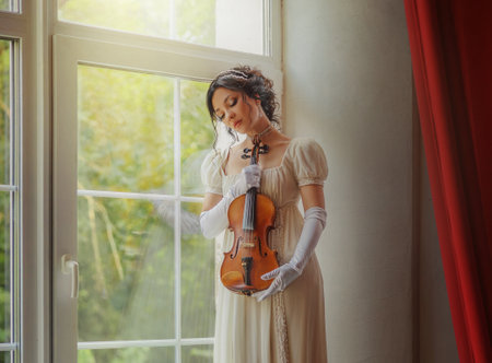 Fashion model woman violinist posing holding in hands wooden violin classic old style. Girl standing on sill window view on green garden light room. Sexy lady beauty face White vintage long dress artの写真素材