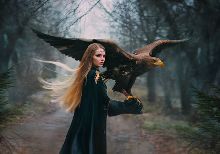 art portrait real person fantasy woman assassin warrior holding white-tailed eagle bird flaping open wings on hand. Elf girl with brown bird walking in forest. Lady queen hunter black cape dark treesの写真素材