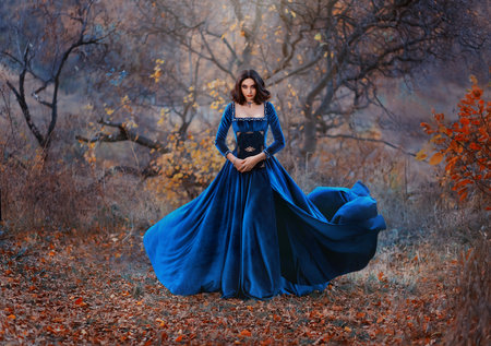Gothic fantasy woman witch holding magic book hands. Long black velvet medieval vintage dress flies wind. Girl conjures. Bright divine light glow sparkles magic circle around lady. Autumn forest tree.の写真素材