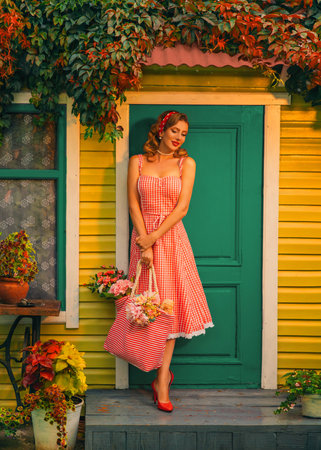 happy retro lady pin up style red lips midi dress smiling face woman standing posing at door of wooden house yard green ivy flowers in pots summer garden walls paneling finishing. Vintage blonde girlの写真素材