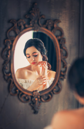 Art photo reflection in antique vintage mirror young beautiful medieval lady woman long hair elegant hairstyle. White victorian old style dress bare shoulders. Fantasy sexy princess looking at mirrorの写真素材