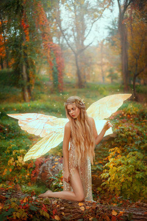 Young fantasy elf woman in image of fairy. Forest pixie girl with golden glowing butterfly wings. Fashion model posing in forest. autumn nature red orange foliage of trees green grass. Lady goddessの写真素材