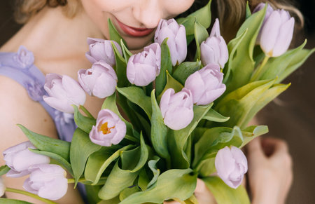 Happy blonde woman holding tulip bouquet purple flowers green leaves white window light. Joyful beauty girl enjoys gift back rear view. Womens day holiday, march 8, spring concept floral greetingの写真素材