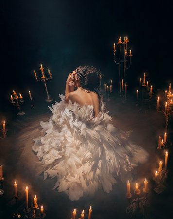 art photo real person girl princess sitting surrounded by candles light flame dark room old style. Fantasy woman bird open bare back rear view black hair. white dress like flower natural featherの写真素材