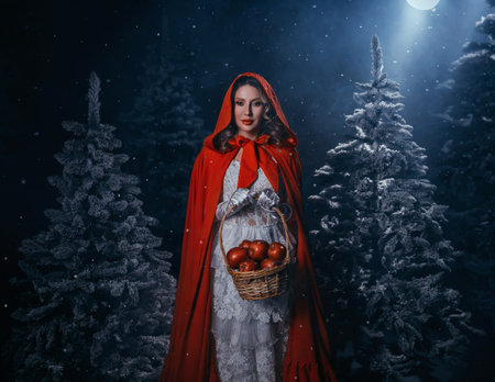 gothic woman walking in fairy tale dark night forest tree winter cold nature snow falling. Fantasy girl hands holding basket of apples. cape white dress red hood on head. moon light fog. Lady queenの写真素材