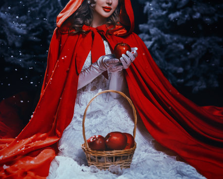 woman queen sits on ground under snowfall in dark night forest tree winter nature snow falling. Fantasy girl beauty cropped face close-up red lips hand holding apple. cape white dress hood on headの写真素材