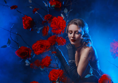 art photo real person sexy fashion model gothic woman posing in fairy tale dark night garden red roses flowers. Fantasy lady beauty face holiday makeup red lips girl black dress. blue moon light fogの写真素材