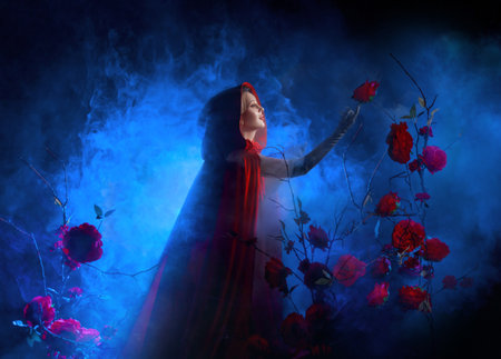 art photo real person gothic woman queen walking in fairy tale dark night garden roses flowers bush tree hands raised to sky. Fantasy lady girl long cape dress red hood on head. blue moon lightの写真素材