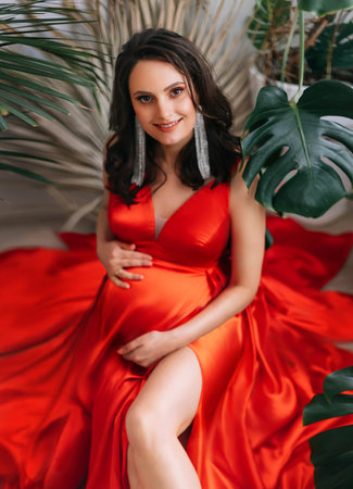 Young happy pregnant woman with smile on face, gently hugs embrace her stomach belly with hands. Red...