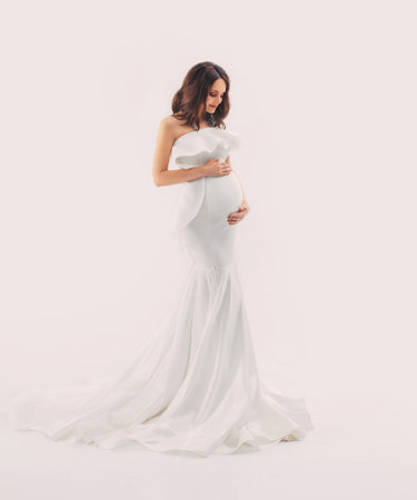 Happy pregnant woman in white dress gently hugs touch her belly with hands. Light beige studio background. Beautiful pregnancy, motherhood, expecting a baby, family health concept. Free space for textの写真素材