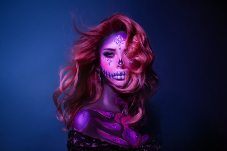 A young beauty woman with long hair in the image of a sugar skull. Hair flying in wind. Halloween carnival party. Close up portrait. A cute sad face in neon colored lightの写真素材