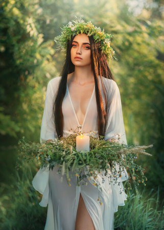Portrait Fantasy Slavic woman holding herbal wreath, burning candle in hands. Divination ritual romantic girl witch sorceress conjures pagan ritual. White silk dress, long black hair. summer natureの写真素材