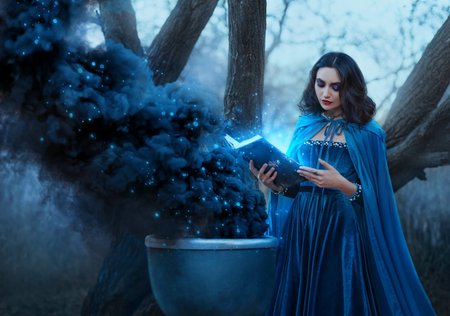 Halloween concept fantasy woman witch conjures, holds book in hands reads spell black magic smoke from boiling vat. Gothic sexy girl. Blue creative design dress cape. Forest trees dark autumn natureの写真素材