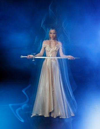 real person sexy face fantasy woman queen holding magic glowing sword in hands. Black dark night room blue moon light fog. Gothic girl warrior with sharp blade shiny dress mixed light art photographyの写真素材