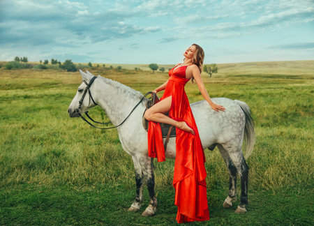 Happy woman sitting on horseback hand stroking white fur mane fashion model posing. girl with horse walking, green grass summer nature blue sky.の写真素材