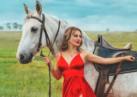 Happy woman hand holding reins white fur mare. fashion model glamour girl posing with horse, green grass summer nature blue sky. lady rider in red long dress fluttering waving in wind blowの写真素材