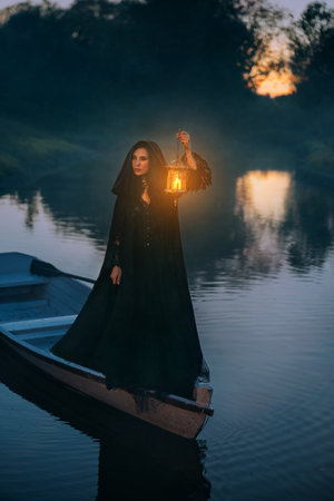 art photo real person fantasy gothic woman floating in boat down river night nature, hand holding lantern warm light. witch girl in ship along lake fog dark forest trees. Black dress cape hood on headの写真素材