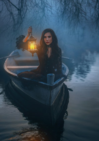 Portrait brunette hair fantasy gothic woman floating in boat down river night nature, hand holding lantern with magic light. Face girl in wooden ship lake fog dark forest trees. Black lace dressの写真素材