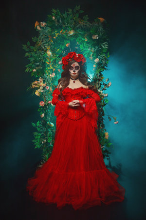 beauty face woman sugar skull make-up eyes lips art paint, long vintage style red dress crown with roses flowers on head. Sexy girl Calavera goddess of death in dark black night garden arch trees fogの写真素材