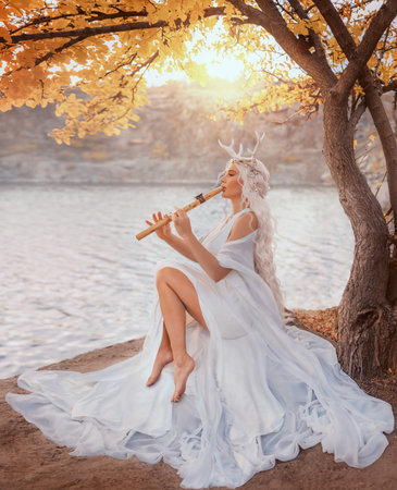 Portrait fantasy woman fairy faun plays a flute, sitting under yellow tree creating magical music. Myth happy beauty face girl fae, long blonde hair white dress deer antlers on head. river rockの写真素材