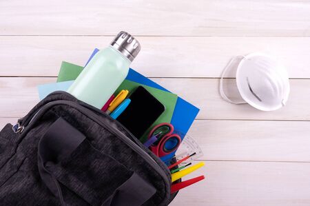 Backpack with school supplies, water bottle, cellphone and face mask on white Wooden background.