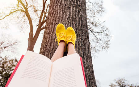 Close-up picture of Female legs leaning on tree wearing yellow sneakers while reading a book. copy space. Selected focus.の写真素材