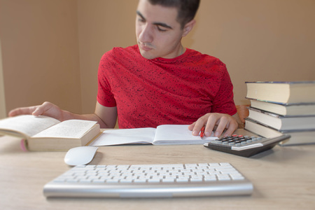 Young man reading book at table in room. Young student write in notebook between books