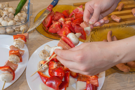 Preparing shashlik for barbecue grill. A hand puts pieces of Champignon, sausages, tomatoes, pepper Skewers on skewer. preparing dinner in a kitchen concept cookingの写真素材