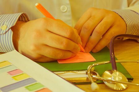 Male doctor with stethoscope sitting at a desk, writing something, prescription or recommendation of...