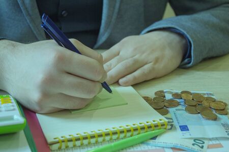 Man is taking notes of saving money in a notebook, with a money jar and a business report on the...