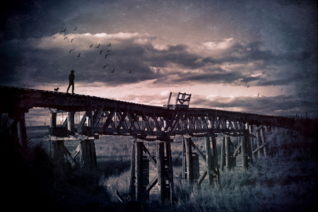 Lone man and his dog wander across an abandoned wooden railway bridge through the countryside. Grunge textured image. Wanderlust, travelling man, wandering soul and mans best friend concepts.の写真素材