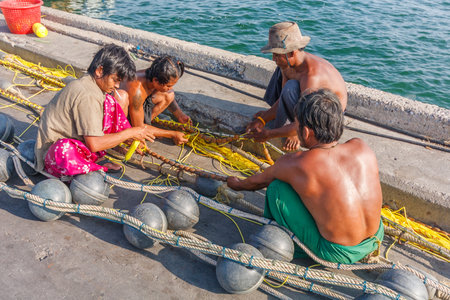 Hua Hin, Thailand - November 17th 2011: Burmese fishermen mending nets. Many Thai fishing boats are...