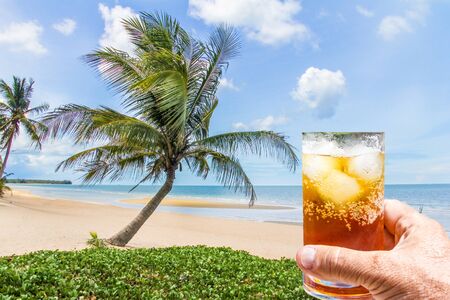 Hand holding a rum and coke cocktail in glass with ice on beach with coconut palm tree, Thailandの写真素材