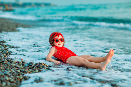 a small, beautiful girl in a red swimsuit and sunglasses, sitting on the beach in summerの写真素材