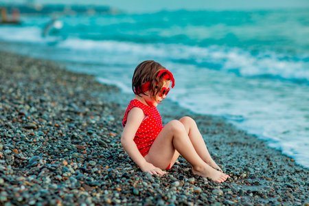 a small, beautiful girl in a red swimsuit and sunglasses, sitting on the beach in summerの写真素材