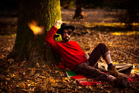 a brutal young man with a beard, wearing a black hat and a red sweater, in the afternoon in the park under an autumn treeの写真素材