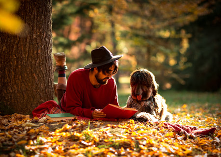 a young man, dark-haired, in a red sweater and hat, lies under a tree with a pet dog, in the afternoon in an autumn parkの写真素材