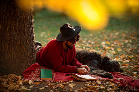 a young man, dark-haired, in a red sweater and hat, lies under a tree with a pet dog, in the afternoon in an autumn parkの写真素材