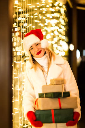portrait of a cheerful girl, in a white fur coat, Santa hat and red mittens, stands in the evening...