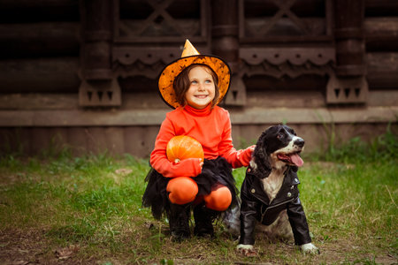 a small, beautiful girl in an orange and black witch costume and a hat, sitting on the porch with a dog in a leather jacket and a pumpkinの写真素材