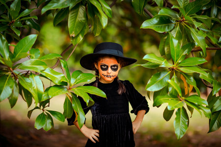 a small, beautiful girl in a black dress and hat, a face in Halloween makeup, stands near a tree in the parkの写真素材