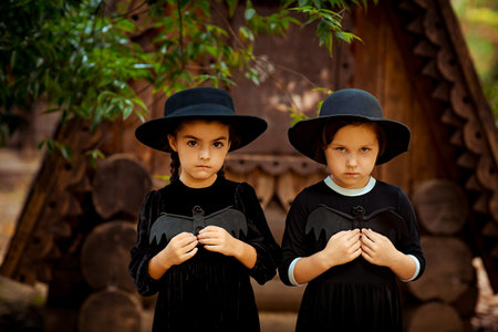 Two little girls in black dresses and hats, standing in the park and holding bats in their hands, for the Halloween holidayの写真素材