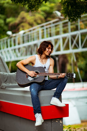 A young, handsome man with long, dark hair, playing an acoustic guitar, on the streets of the city, in a white T-shirt and jeansの写真素材