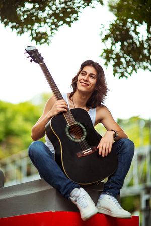A young, handsome man with long, dark hair, playing an acoustic guitar, on the streets of the city, in a white T-shirt and jeansの写真素材
