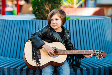 A beautiful little girl, in rock style, in a leather jacket and jeans, stands on a summer stage, and plays an acoustic guitarの写真素材