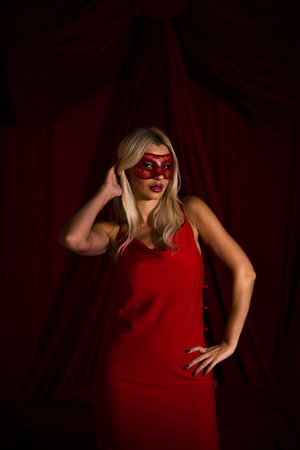 A blonde woman in a red, long dress and a masquerade mask, in a studio on a red backgroundの写真素材