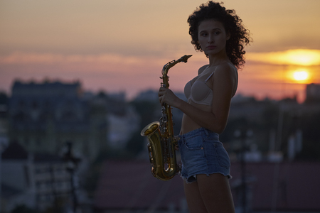 Young curly girl with sax on the roof at sunsetの写真素材